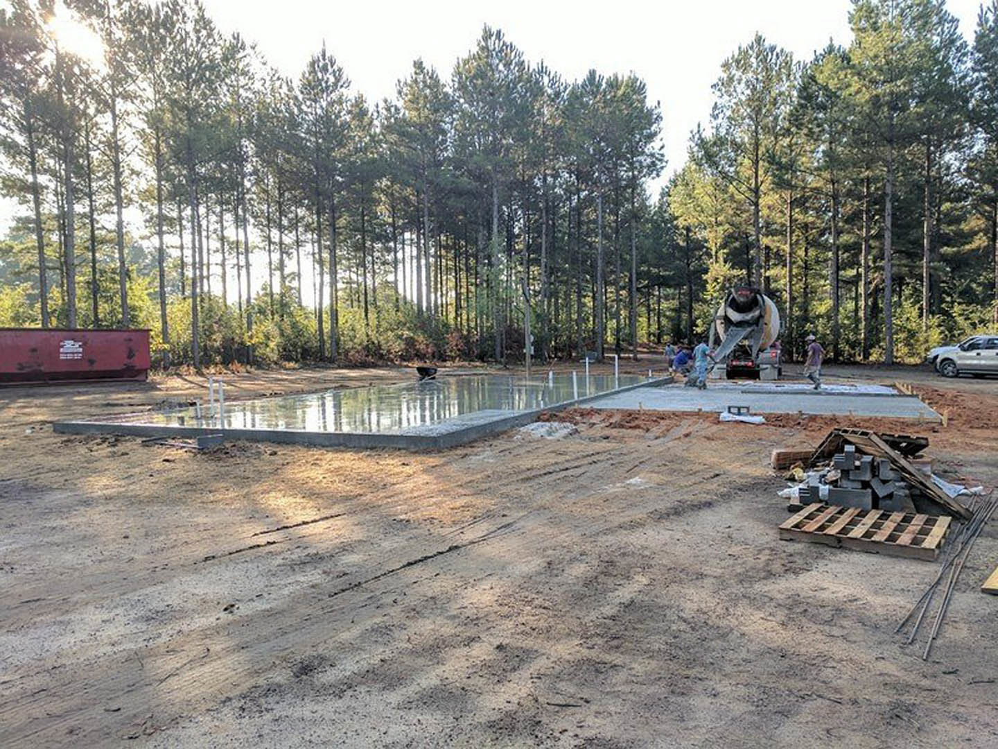 Concrete slab foundation with cement mixer beside an in-ground pool, surrounded by dirt and mature trees