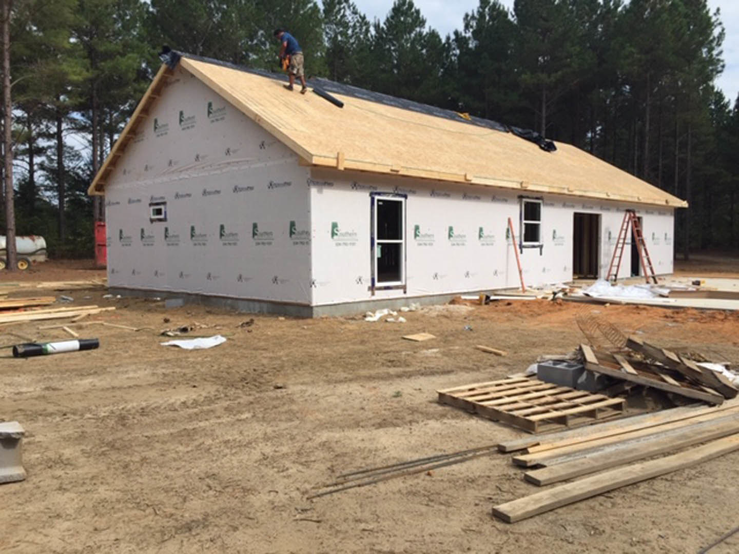 Blurry view of a man standing on a roof above a white-walled house with a white-framed window, surrounded by trees and sky