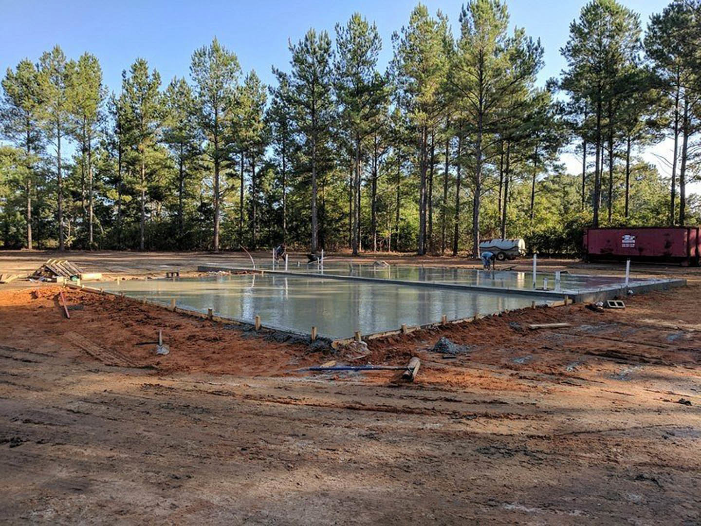 Partially built pool surrounded by concrete slab, construction materials, and mature trees under open sky