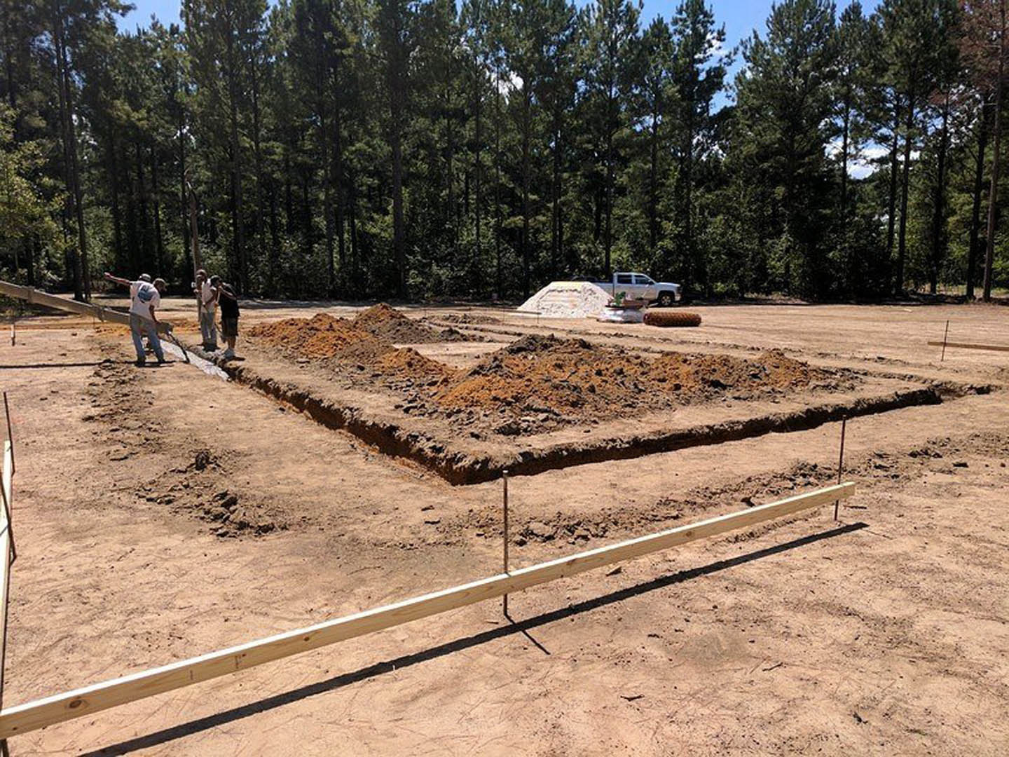 Workers assembling wooden beams on dirt foundation, surrounded by trees and soil, with stone materials visible in foreground