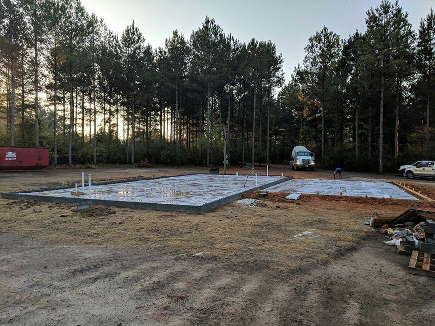 White truck parked on dirt construction site beside a red utility box, metal pool railing, and cluster of tall trees under a clear sky