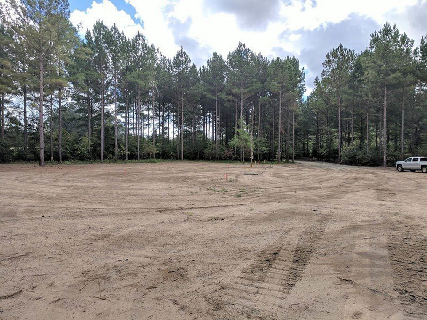 Dirt field with tire tracks bordered by dense trees and a tall leafy tree, white truck parked on nearby road under cloudy sky