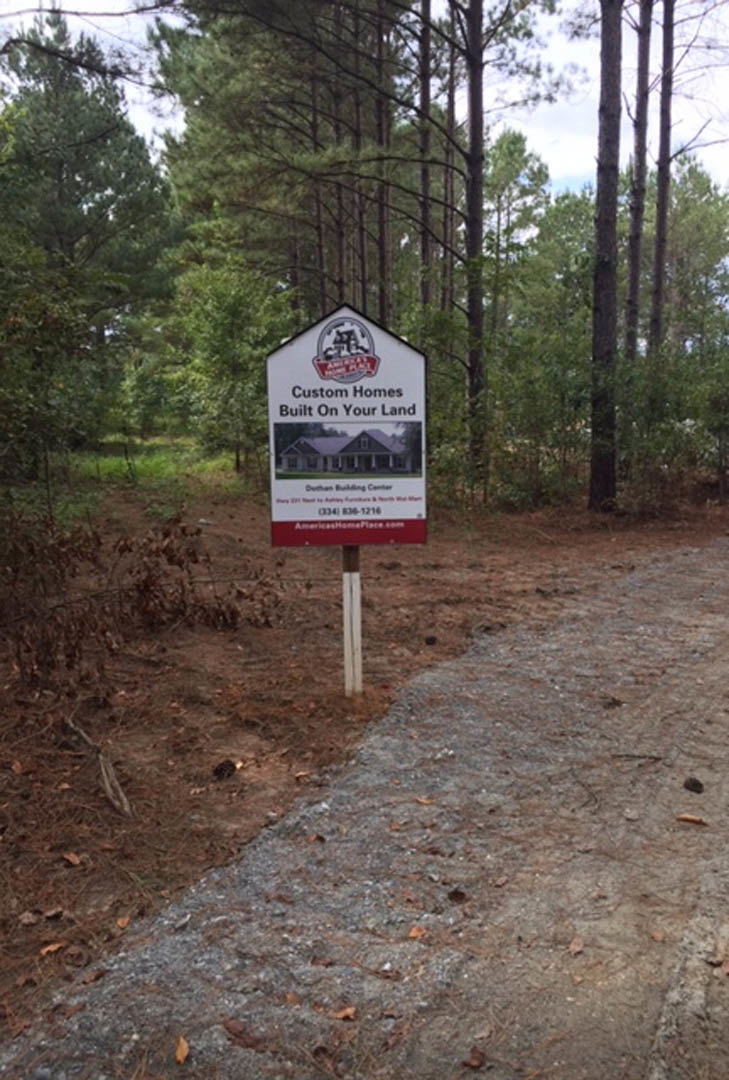 Wooden sign with house illustration stands beside a dirt road surrounded by tall trees and dense forest vegetation.