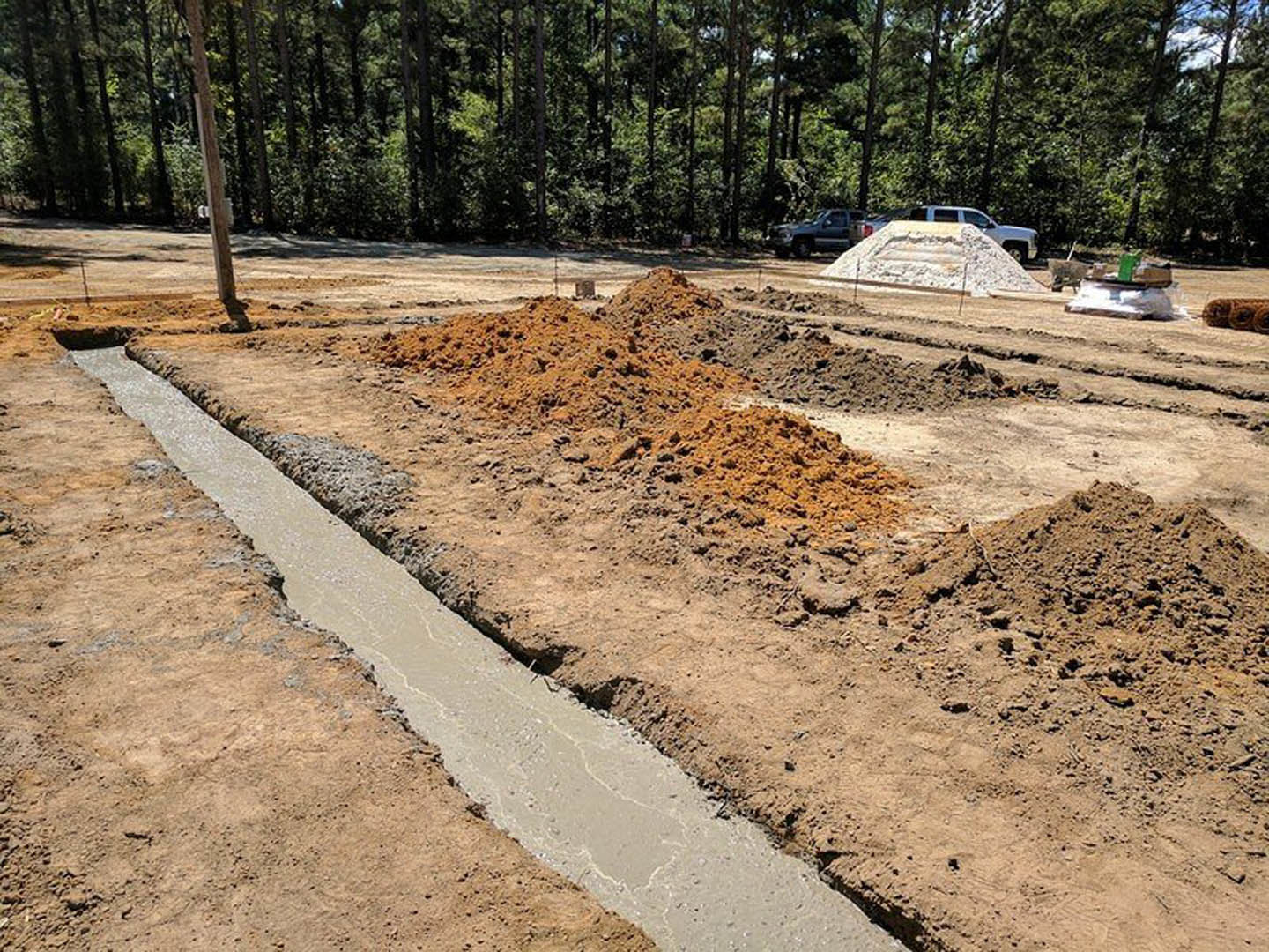 Dirt construction site with open trench, wooden pole bearing sign, forested tree line in background, parked car near stone monument, green utility box partially submerged in water