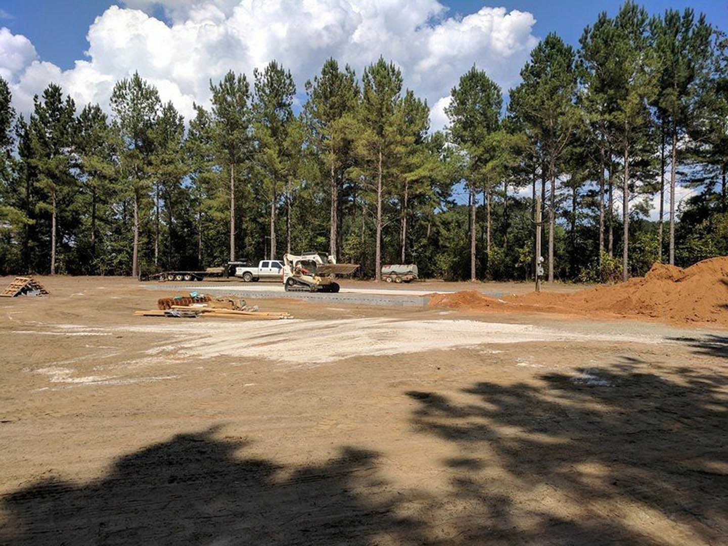 Dirt construction site bordered by mature trees, scattered vehicles parked on sandy ground, tree shadows cast across the site, partly cloudy sky overhead