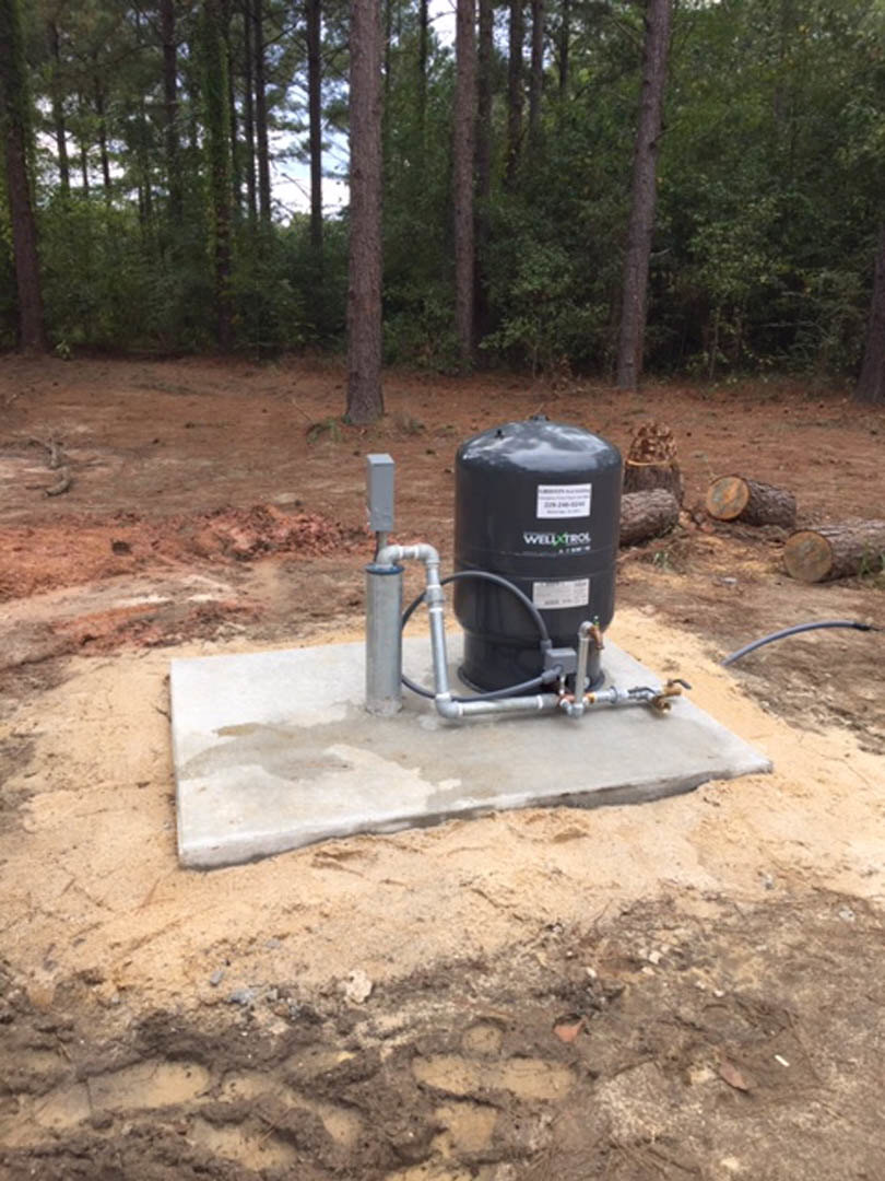 Black water tank with attached hose and pipes sits on a concrete slab surrounded by dirt, with trees and logs in the background.