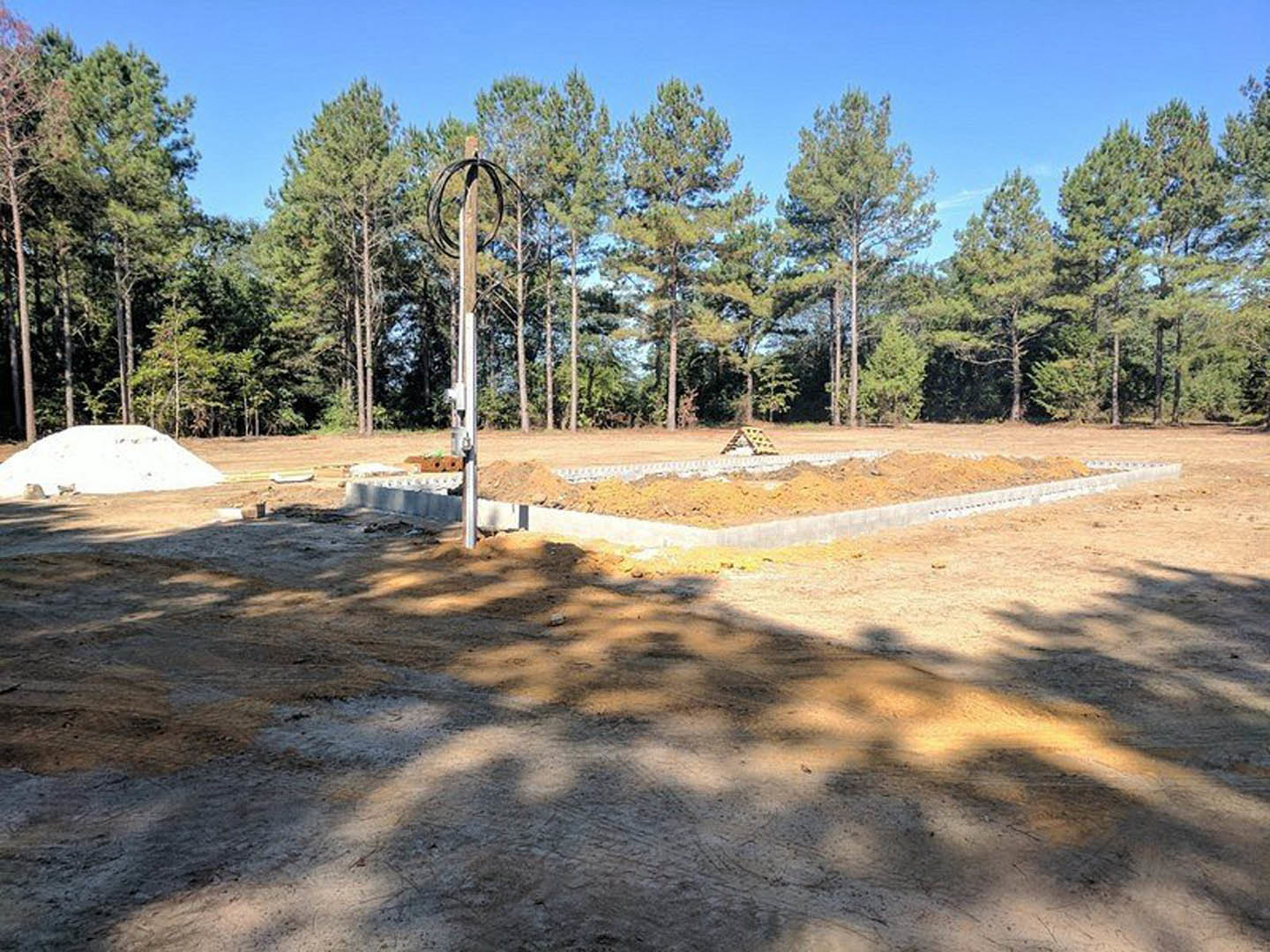 Dirt construction site bordered by tall green trees, white car parked on bare ground, white pillar and post near telephone pole with overhead wire, sunlight casting tree shadows