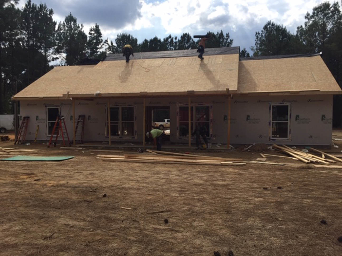 Crew installing shingles on pitched roof of a two-story house with white siding, surrounded by trees under cloudy sky