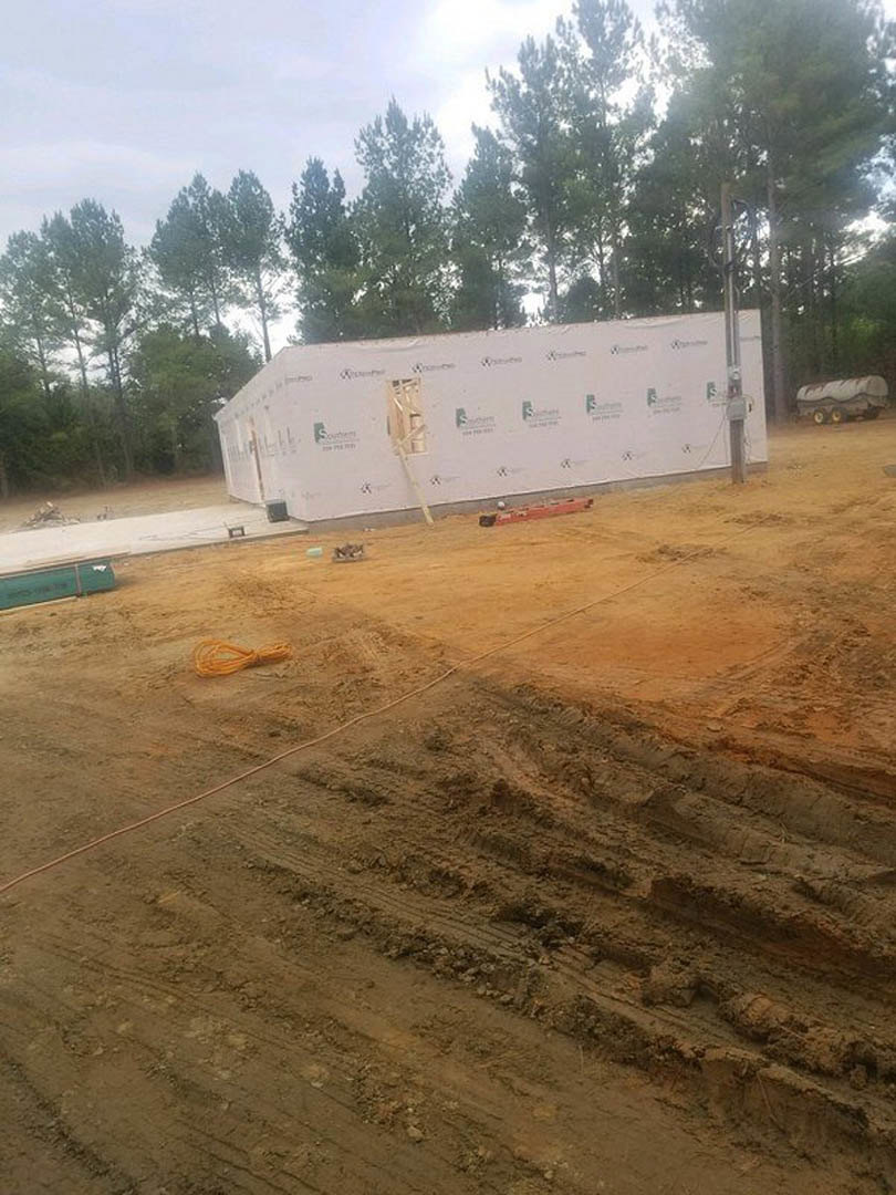 Wood-framed custom home under construction on a dirt lot, surrounded by trees, construction signage, green storage container, and parked truck