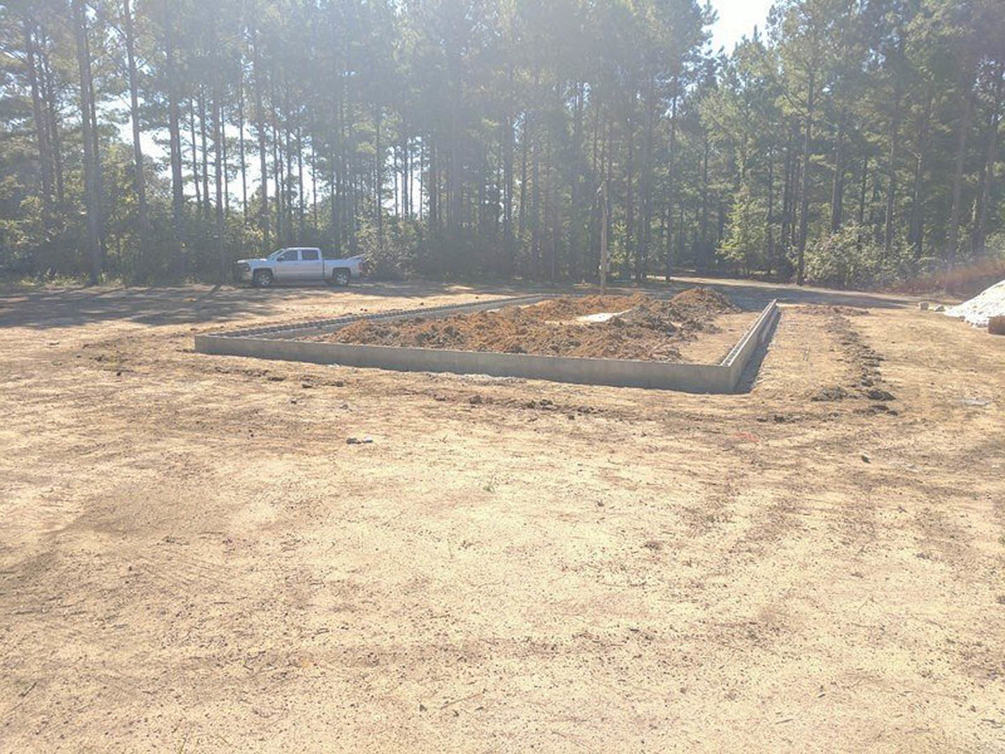 White pickup truck parked on a dirt lot surrounded by soil piles, sparse grass, and a backdrop of leafy trees under a pale sky.
