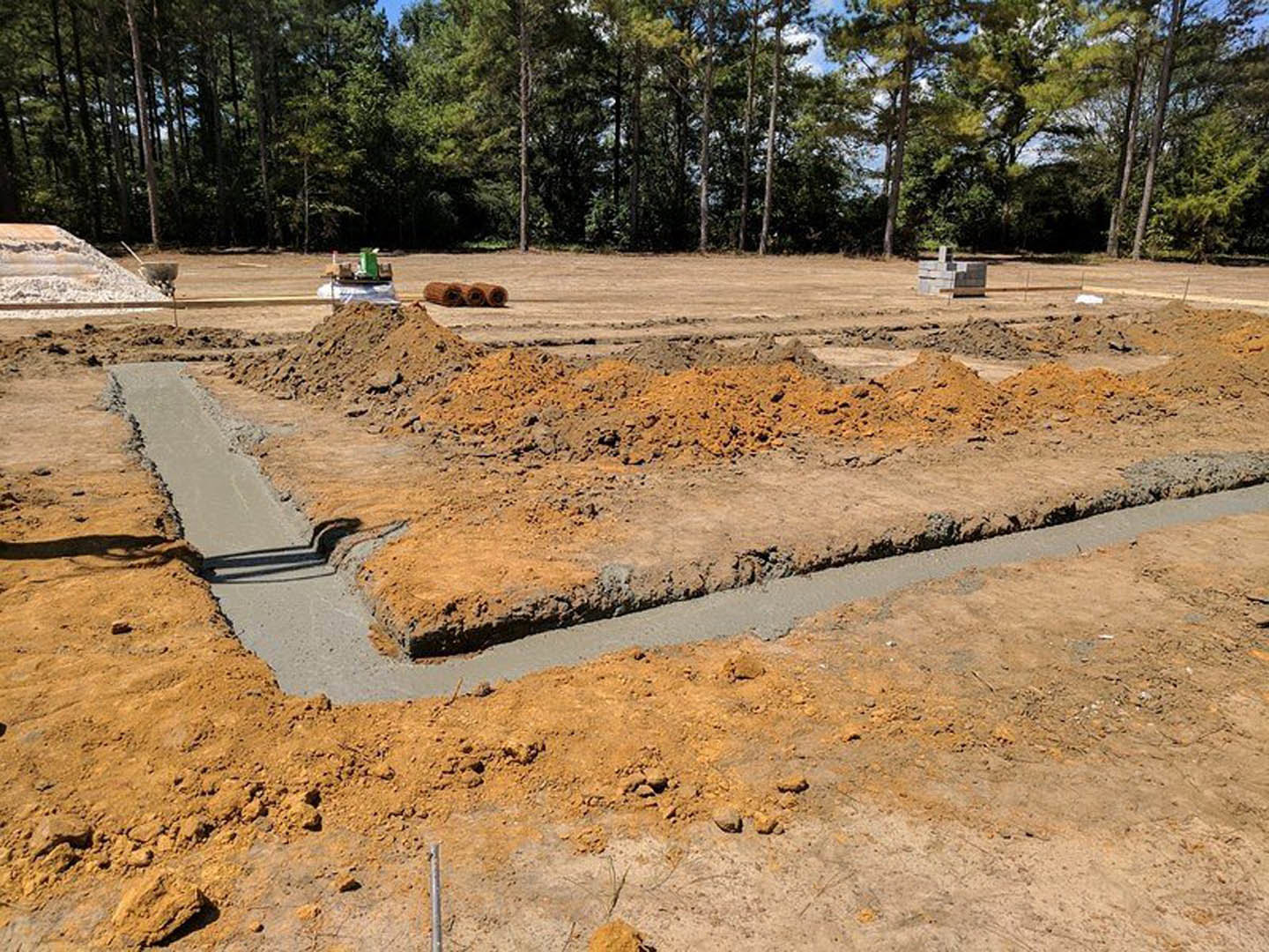 Dirt-filled construction site with exposed soil, scattered gravel, rolls of hay, and surrounding trees under a partly cloudy sky