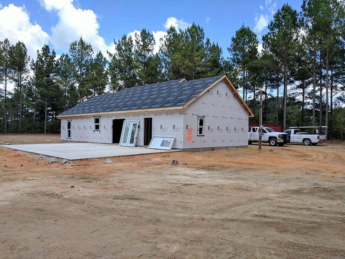 Framed house under construction with exposed wood, open doorway, dirt lot, white truck parked beside utility pole, trees in background, cloudy sky overhead