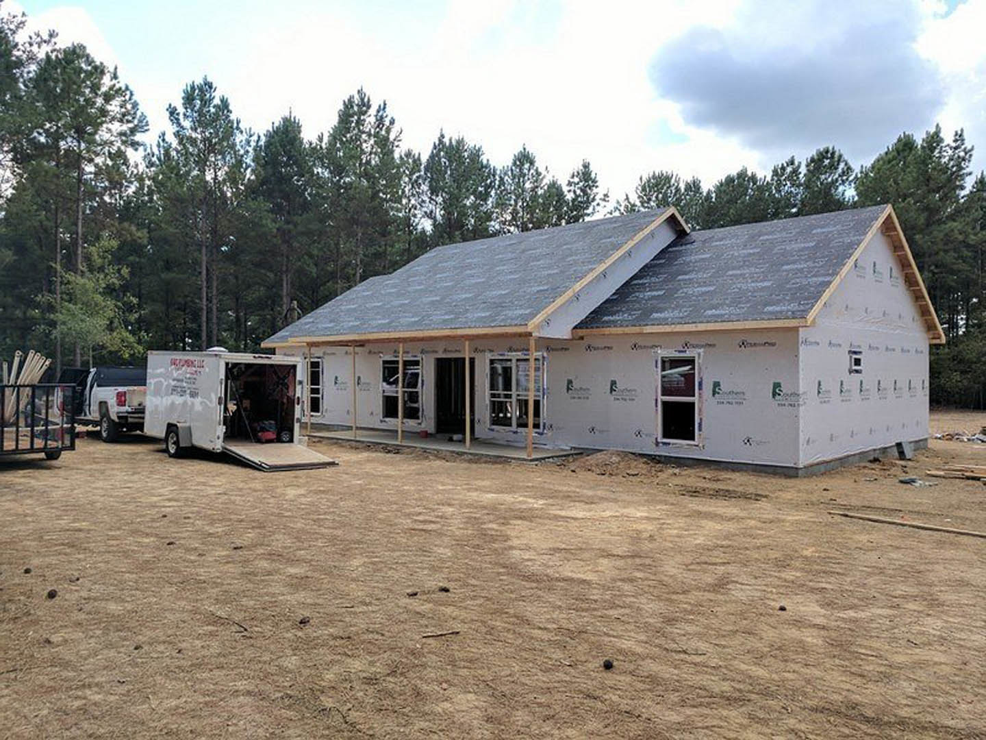 Partially built house with exposed framing and roof, white pickup truck parked on dirt lot, open truck door, unfinished window, cloudy sky, trees in background