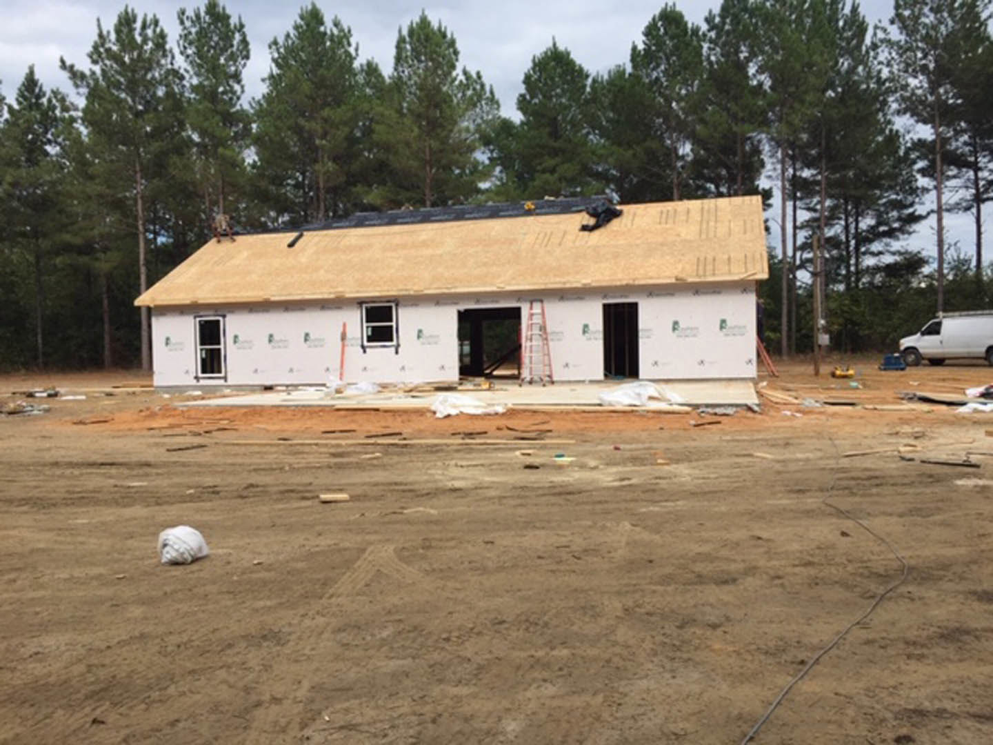 White house under construction with exposed framing, dirt lot in foreground, white van parked with door open, leafy trees in background