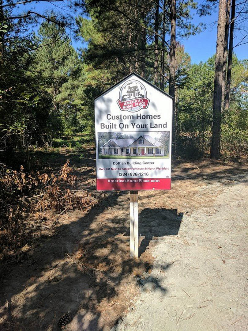 Wooden sign with a house illustration mounted on a white fence post, surrounded by dense trees and forest floor