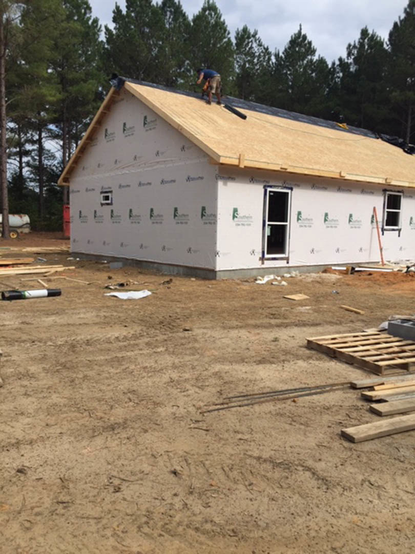 Man in blue shirt standing on unfinished roof, wooden pallets on ground, square-paned window, exposed lumber, trees and sky in background