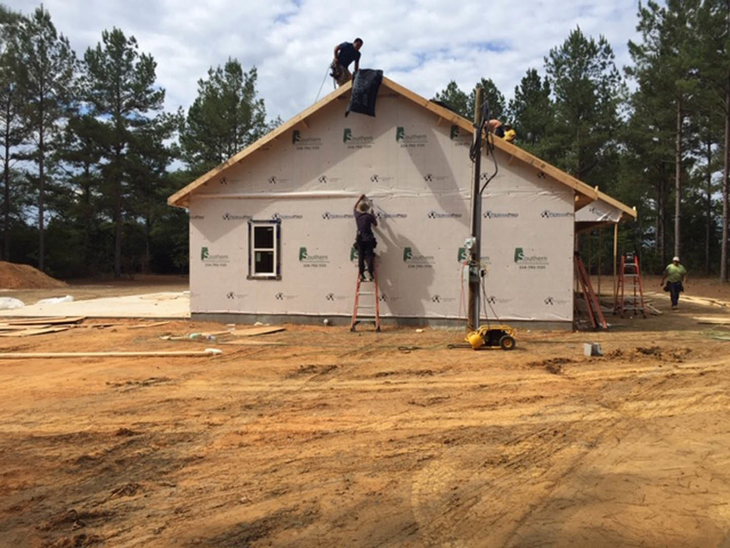 Two men on ladders working on the exterior of a house with white siding, visible roofline, dirt yard, and square window panes.