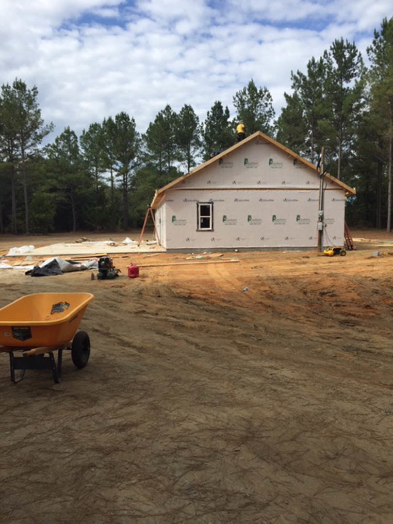 Wood-framed house under construction with exposed beams, wheelbarrow on dirt ground, scattered construction materials, and leafy trees in the background under a partly cloudy sky