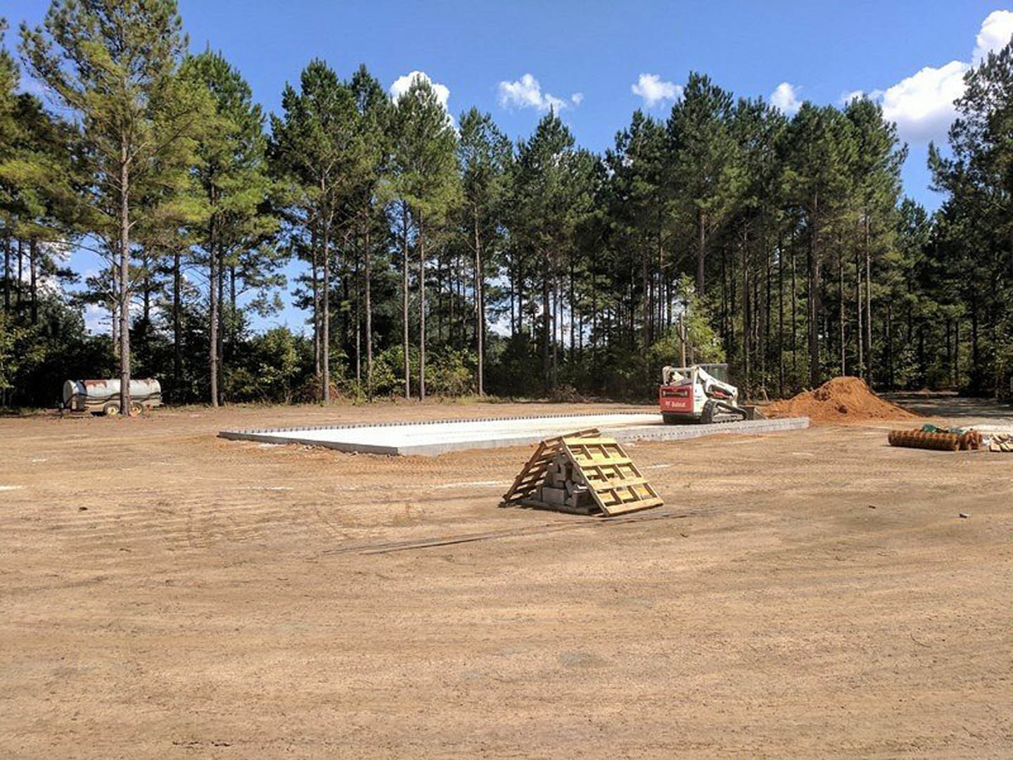 Bulldozer parked on dirt construction site beside stacked wooden pallet, surrounded by soil, grass, and trees under cloudy sky