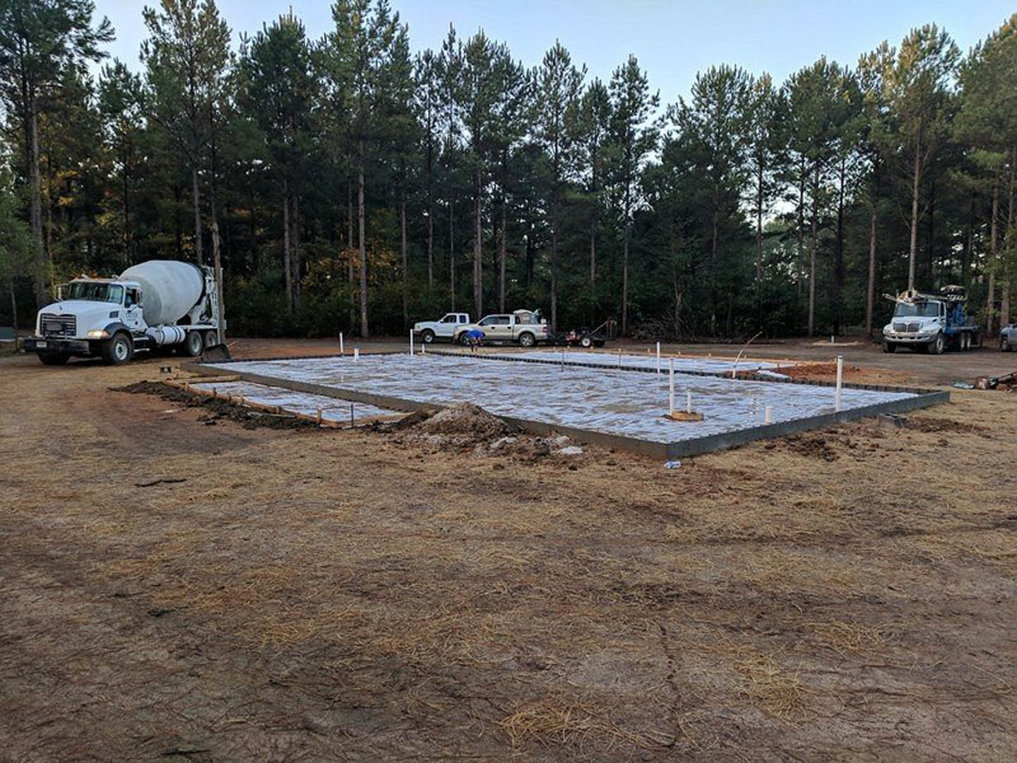 Concrete mixer truck and several vehicles parked on dirt construction site, surrounded by trees and grassy areas, with concrete slab and poles visible in foreground