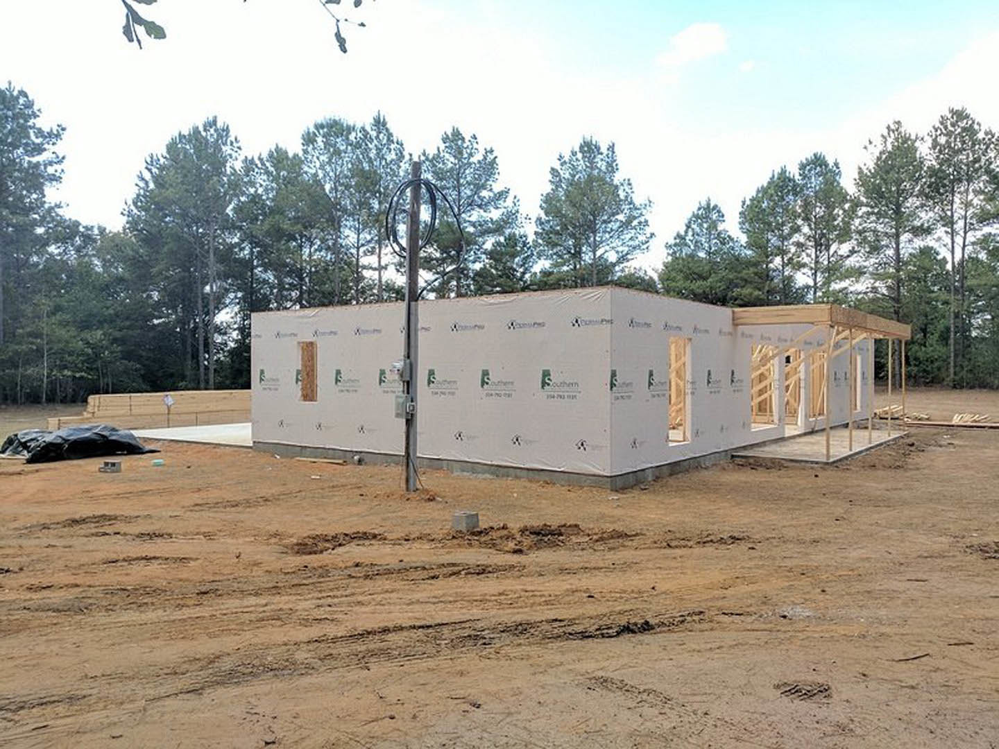Wood-framed house under construction on dirt lot, surrounded by trees, street light, and cloudy sky