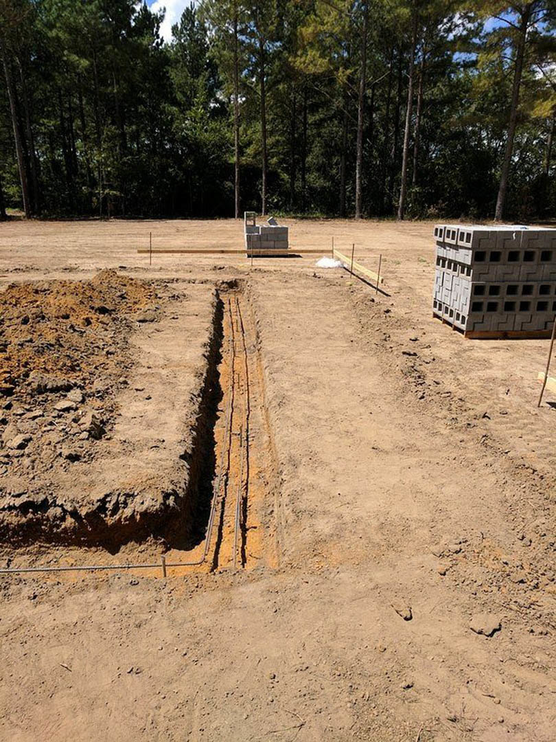 Dirt construction site with stacked cement blocks, scattered bricks, tall trees in background, and partially visible fence