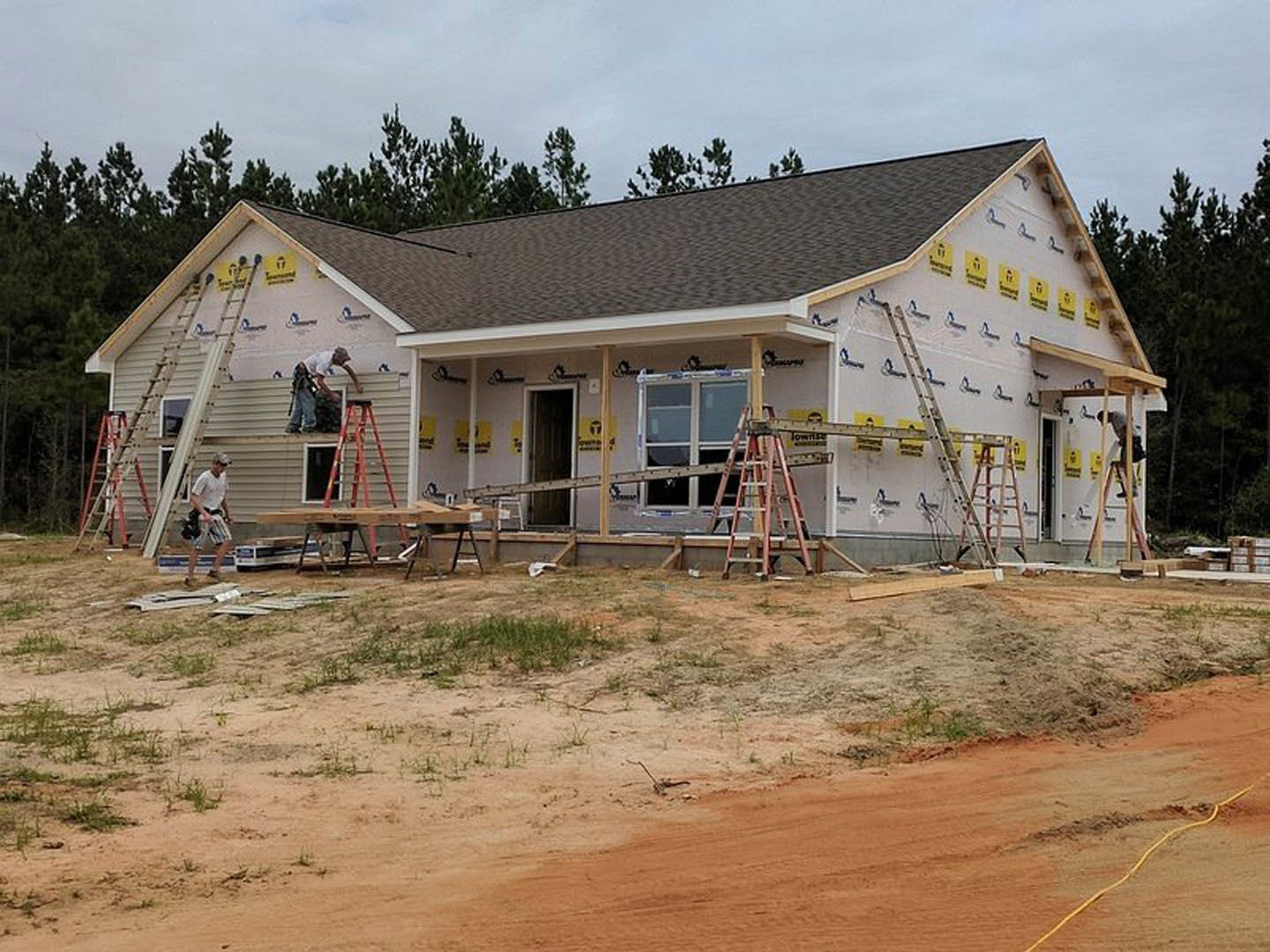 Partially constructed house with exposed framing, multiple ladders leaning against exterior walls, unfinished windows, and construction materials scattered on bare ground