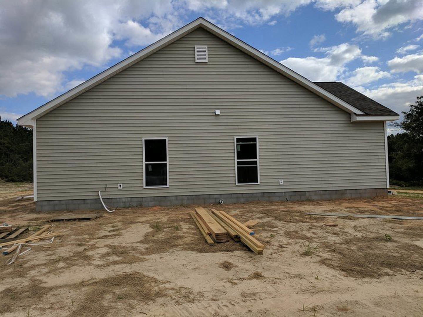 Partially built house with exposed wood framing, white-framed window, wall vent, and pile of wood planks on the ground