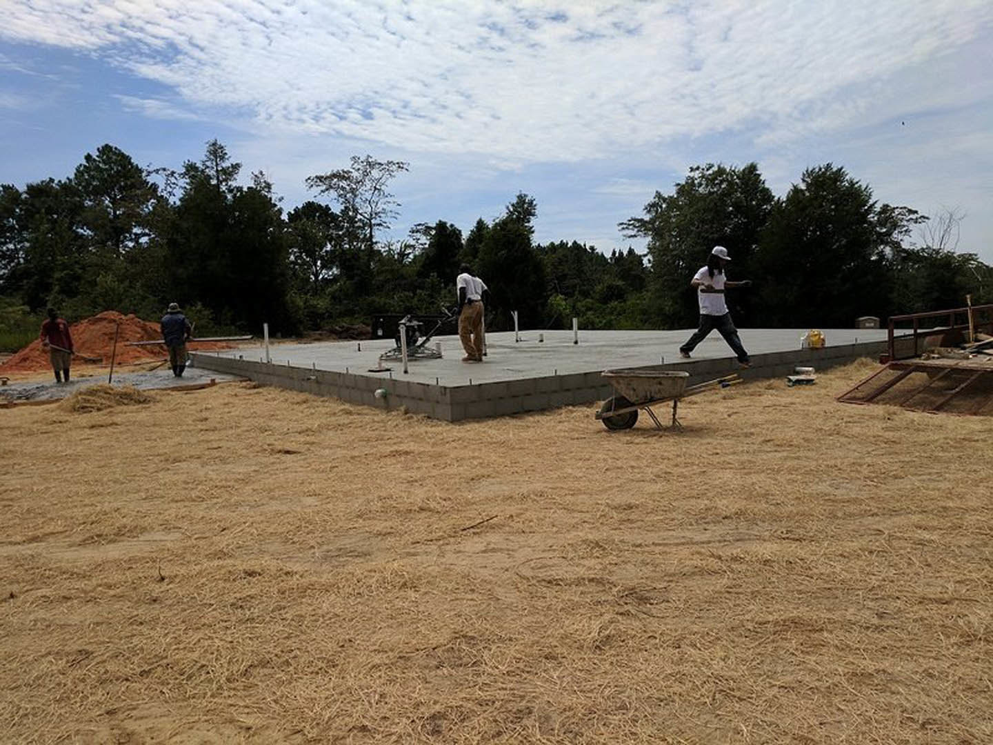 Men building a custom home on a dirt lot with wheelbarrow, concrete blocks, and construction materials; trees and cloudy sky in background