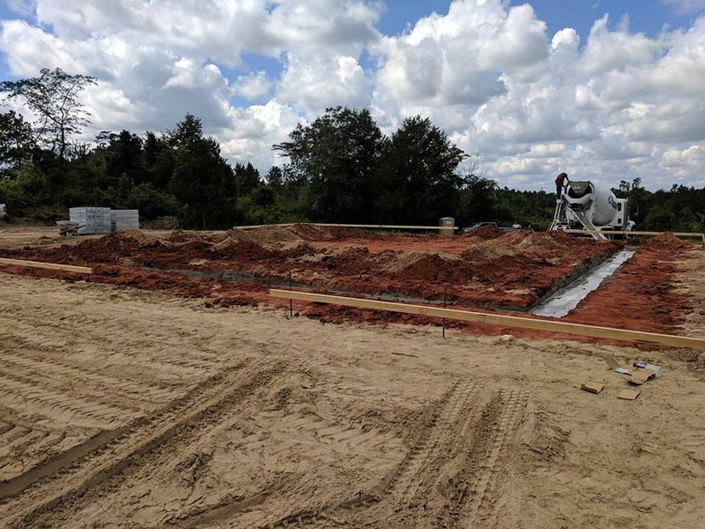 Construction site with exposed soil, tire tracks, scattered wooden beams, cement mixer, worker on ladder, and surrounding trees under cloudy sky