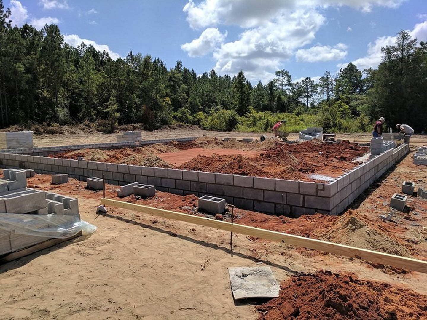 Bricks stacked beside a dirt foundation, wooden boards and fencing surrounding the residential construction site, trees and clouds visible in the background