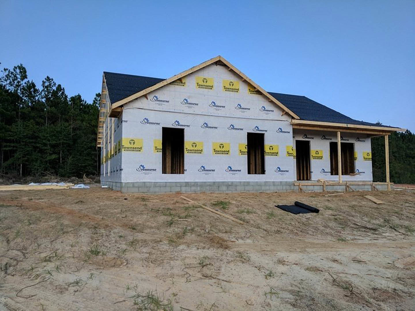 Partially built house with exposed framing and black door, surrounded by dirt field with scattered grass and sticks, yellow and blue construction signs, and trees in the background