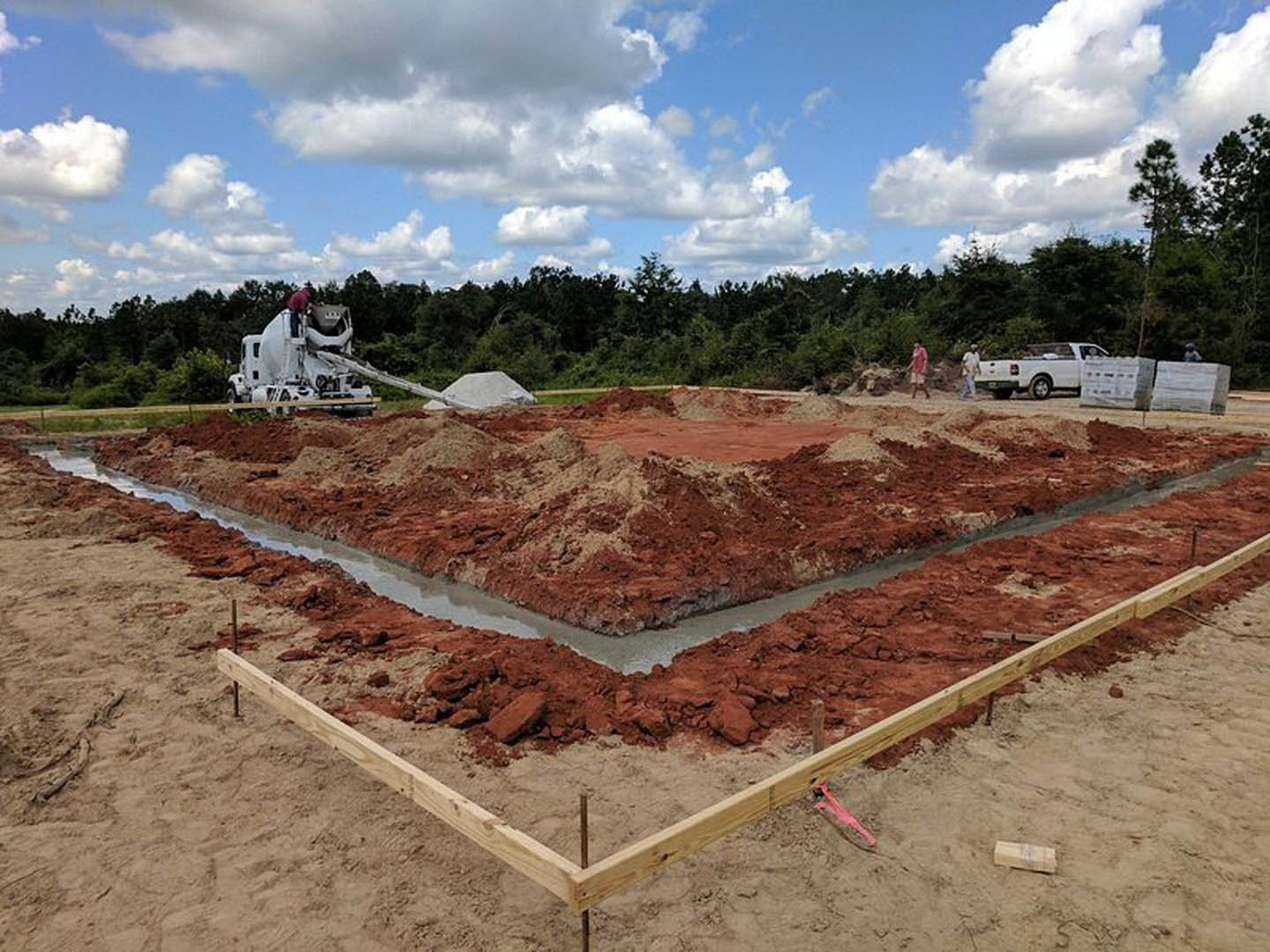 Construction site with dirt ground, wooden fence, white cement mixer truck, man standing on truck, blue sky with scattered clouds