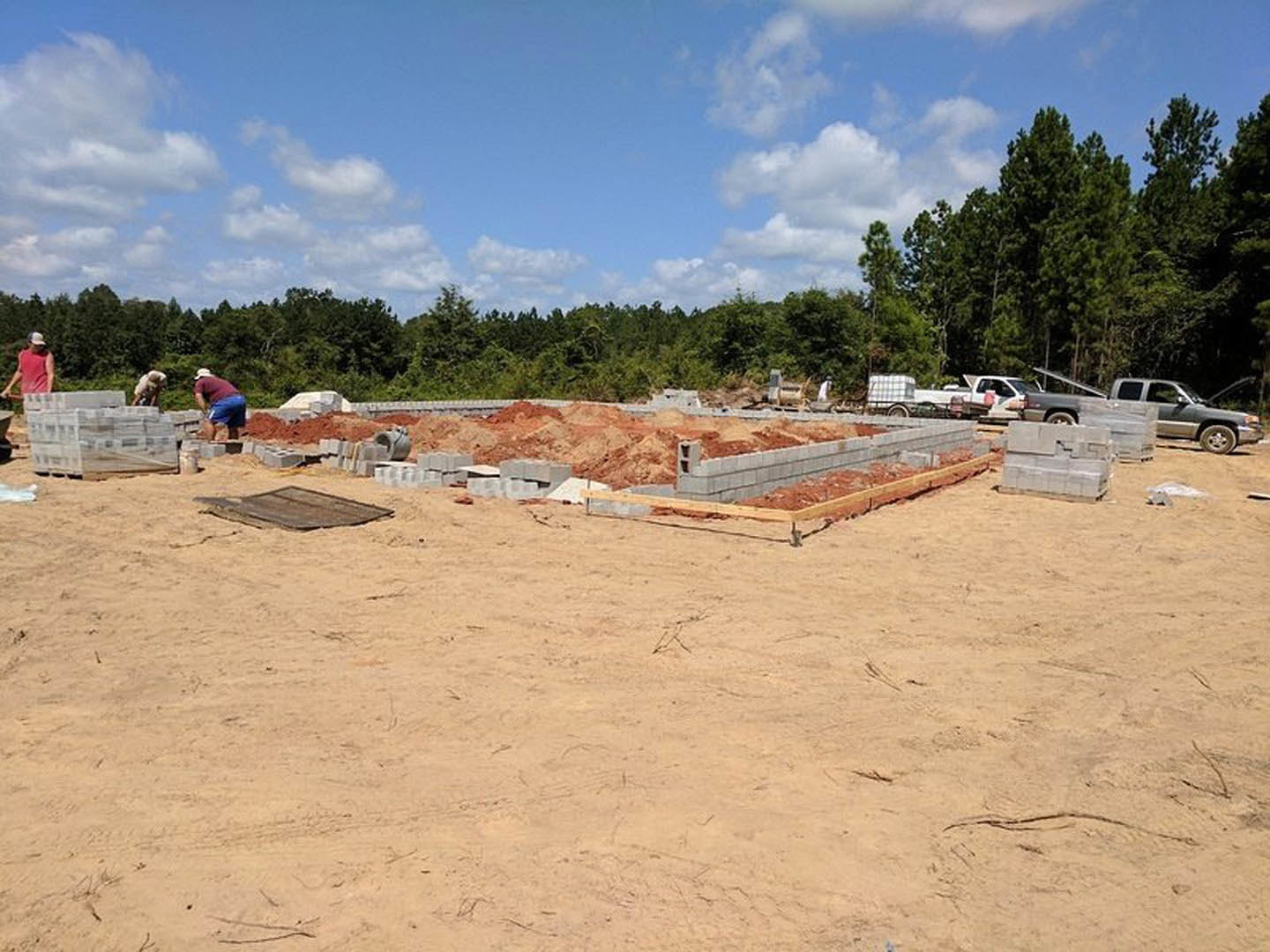 Partially built custom home on a construction site with exposed cement blocks, sandy soil, scattered tools, and workers in red shirts under a blue sky with clouds.