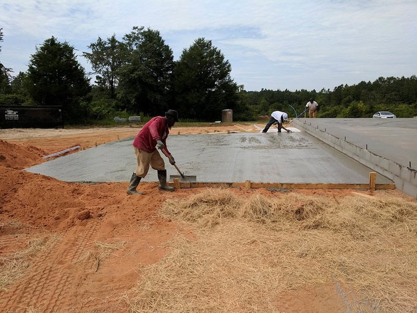 Crew leveling freshly poured concrete slab outdoors, men using shovels and tools, soil and trees visible in background, close-up of work boots and construction materials.