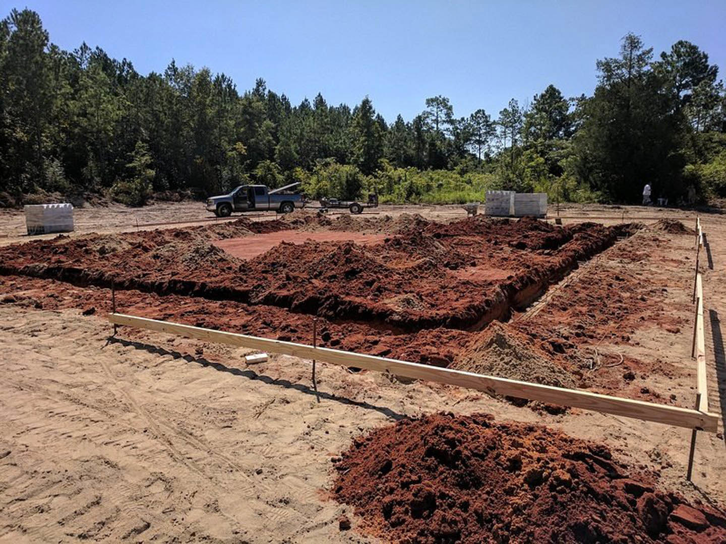 Construction site with exposed soil, scattered wooden beams, open truck door, piles of dirt, surrounding trees, and clear blue sky