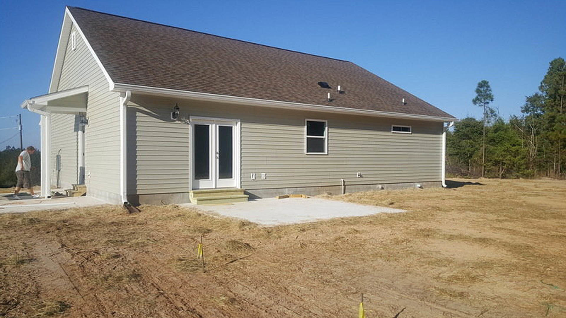 Partially built house with exposed framing, white siding, and visible door and windows, surrounded by dirt lot and scattered trees under a clear sky