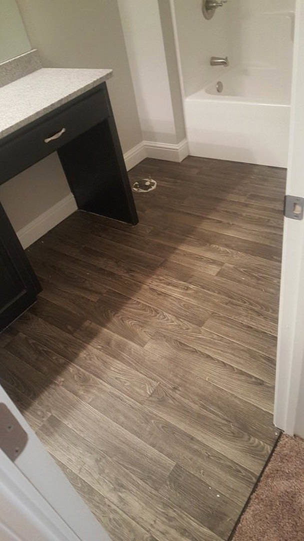 Black desk on wood flooring beside white door, countertop with black drawer, blurred person’s head in background, close-up of carpet and desk, indoor space with tile, sink, and