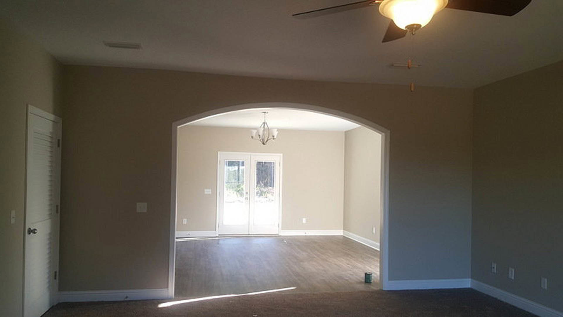 Living room with wooden flooring and white trim, ceiling fan and light fixture, double glass doors, white wall switch, plaster walls