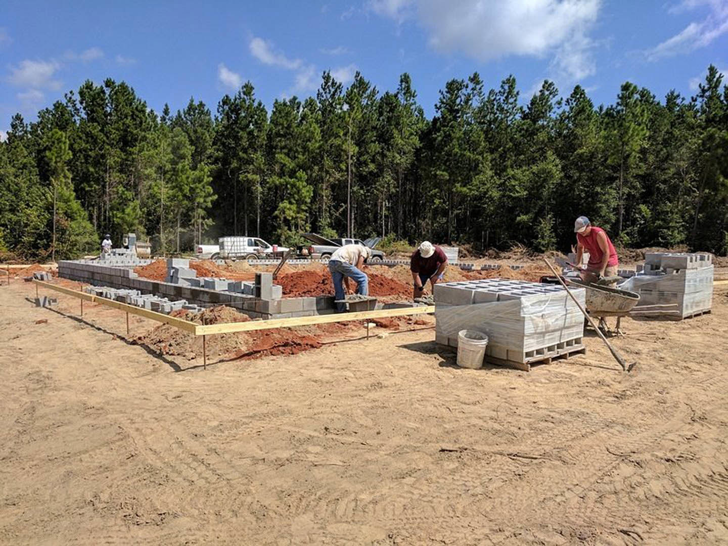 Several men building a custom home foundation outdoors, surrounded by soil, construction materials, and trees under a partly cloudy sky