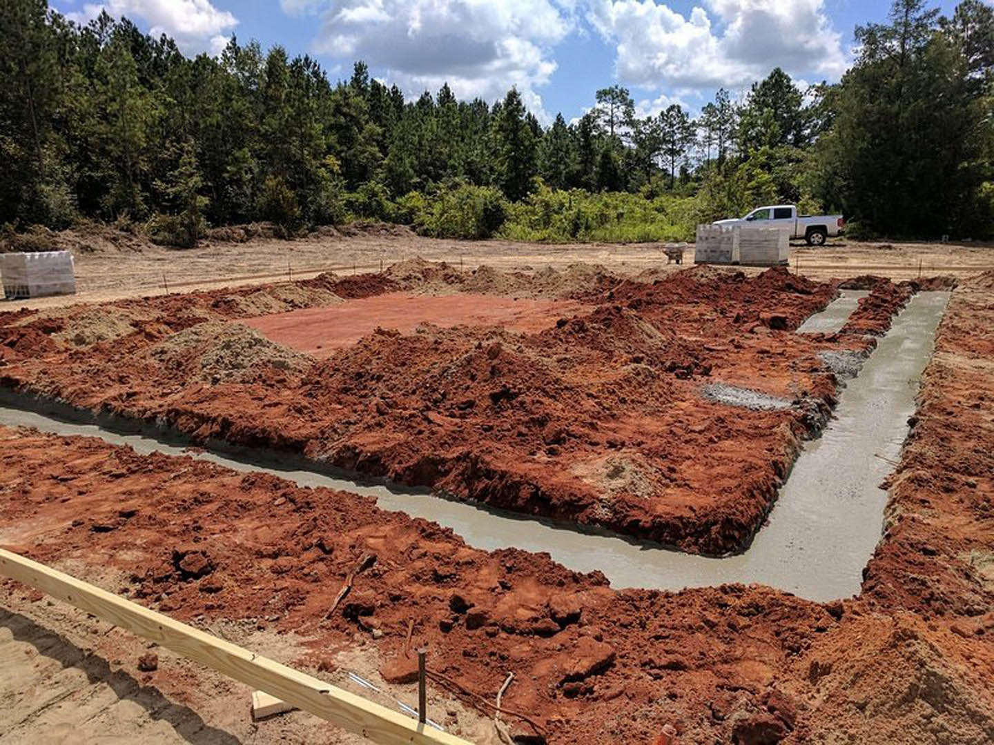 Wooden beam resting on dirt at a residential construction site, white truck parked nearby, white brick wall with metal grate, surrounding trees and forest, cloudy sky overhead.