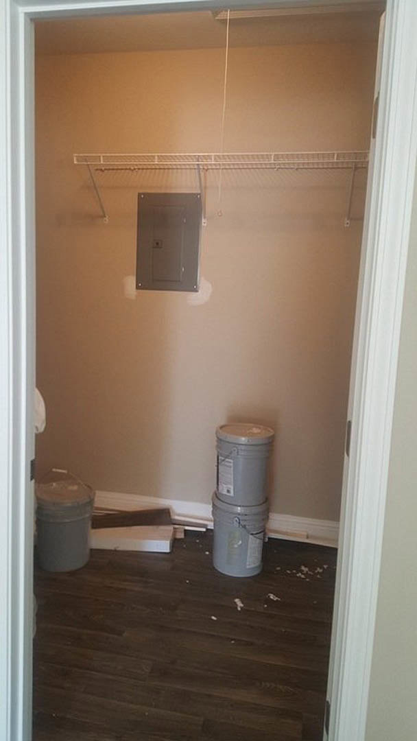 Dark wood flooring in a bathroom under renovation, white door with light switch, grey barrel with lid, grey cylinder with white label, white metal shelf holding a black box