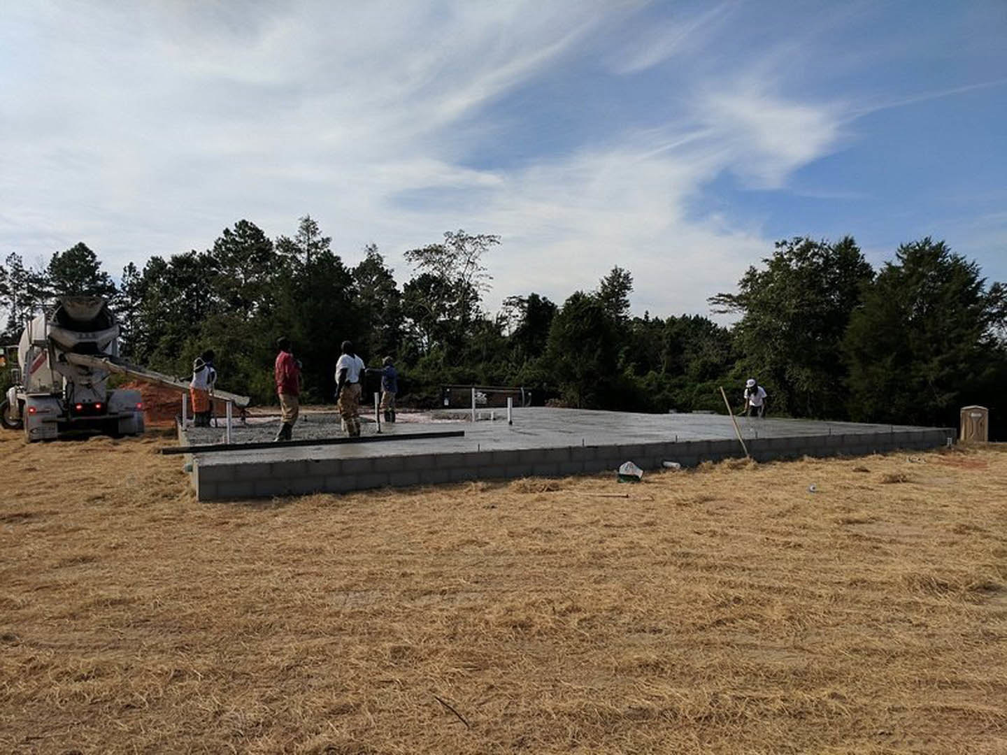 Group of people gathered on a newly poured concrete foundation, brick wall and grassy field in background, construction materials and concrete mixer truck nearby under blue sky
