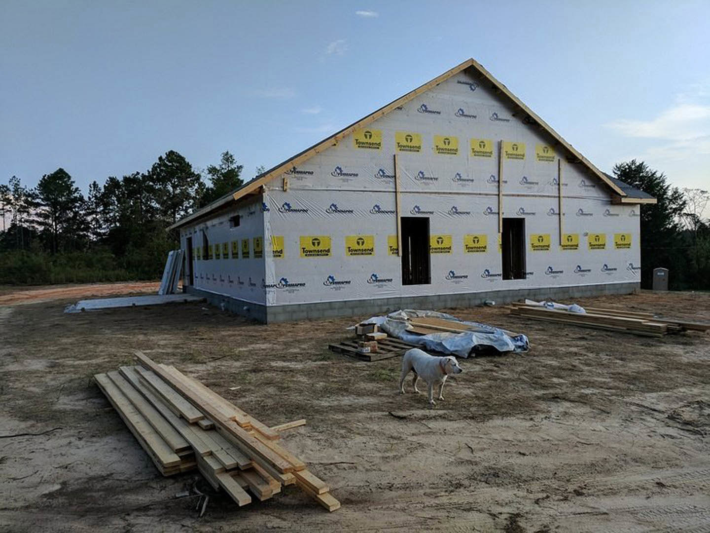 White dog standing on dirt in front of a partially constructed house with exposed wood framing, yellow and black construction signs, and a pile of lumber nearby