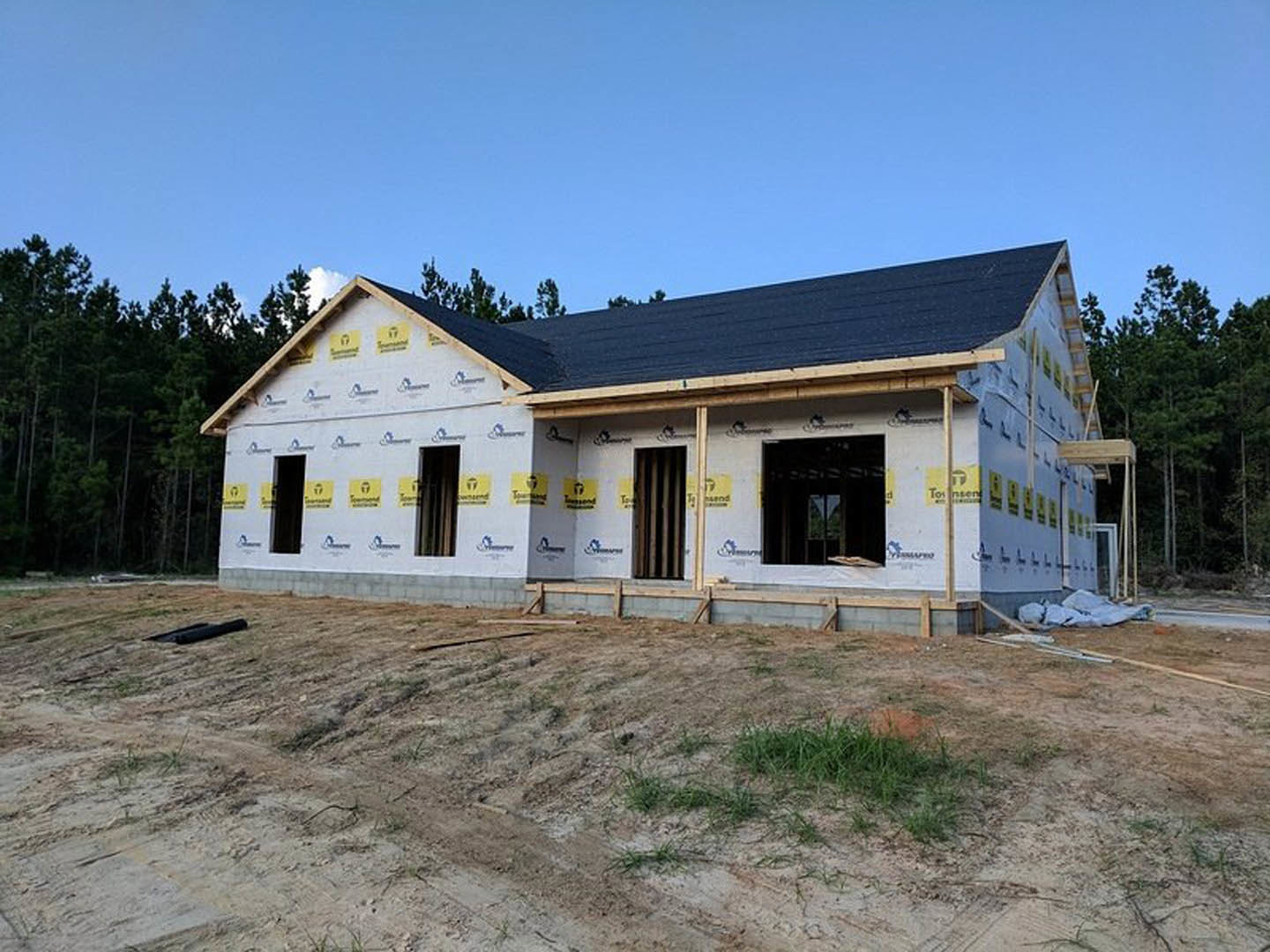 Framed house under construction with black roof, exposed plywood walls, dirt and grass in foreground, mature trees in background