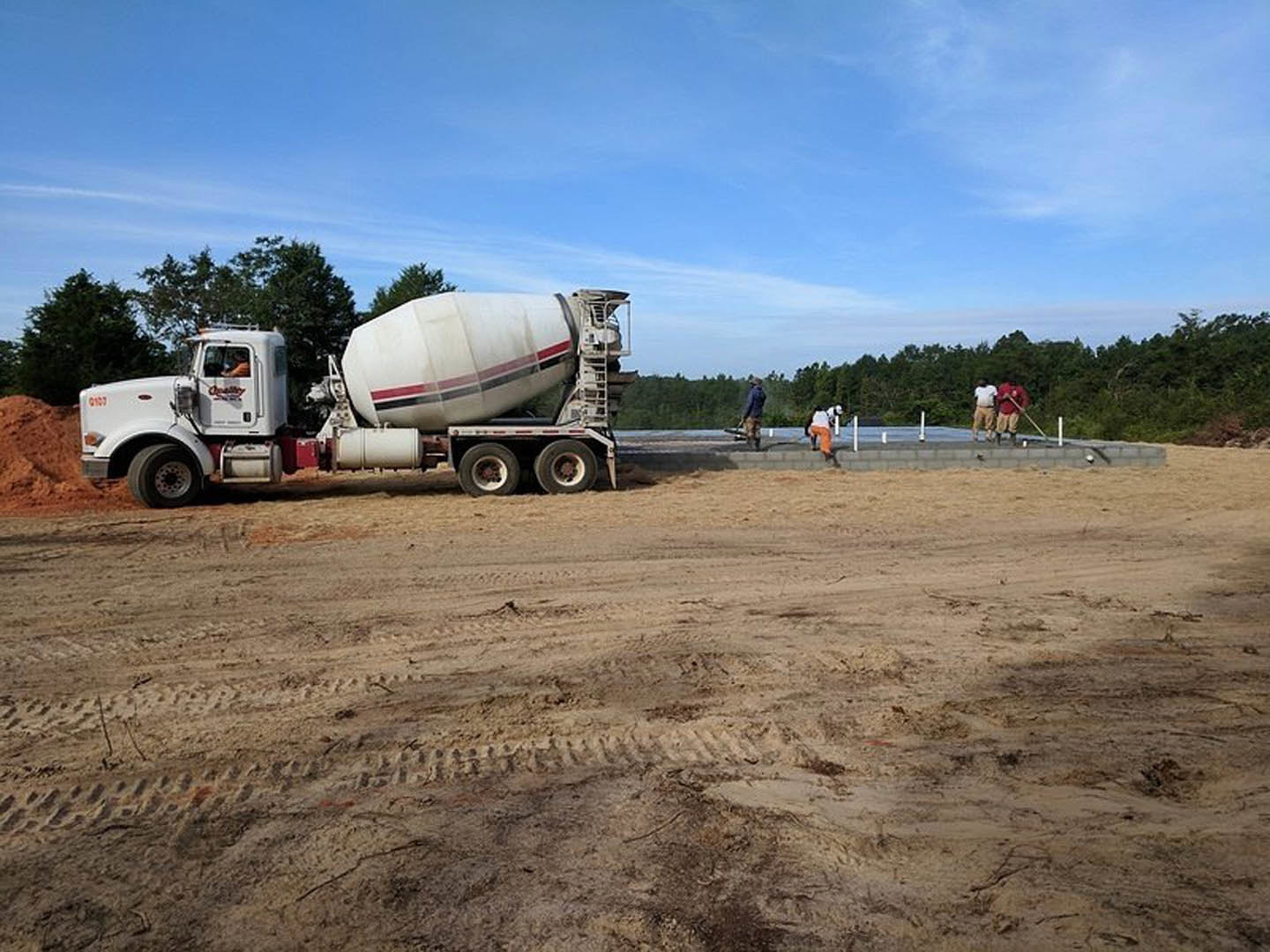 Cement mixer truck with red stripe parked on dirt driveway, tire tracks visible, surrounded by trees and open sky