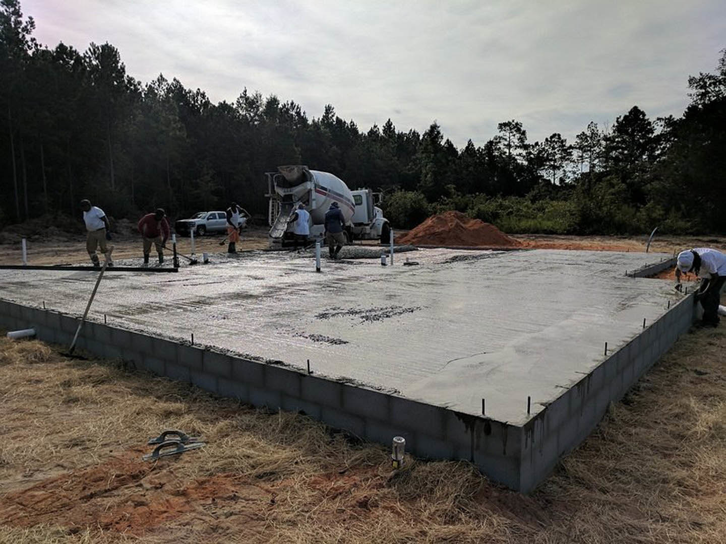 Workers pouring and leveling concrete slab outdoors, surrounded by construction materials and trees under a cloudy sky