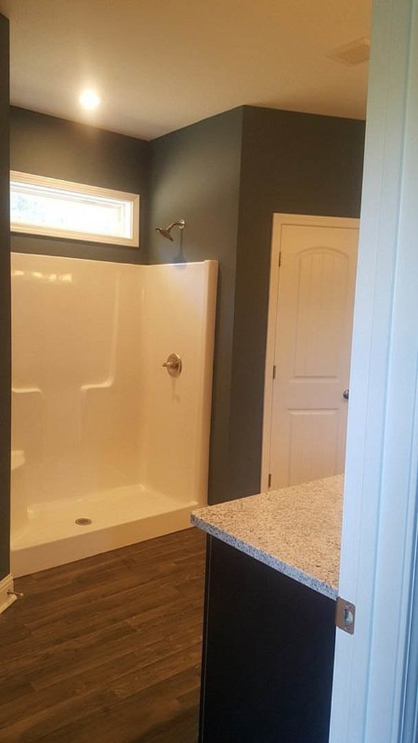 Modern bathroom featuring a glass-enclosed shower with white tile walls, black vanity with white countertop, wood flooring, window with overhead light, and white door with silver