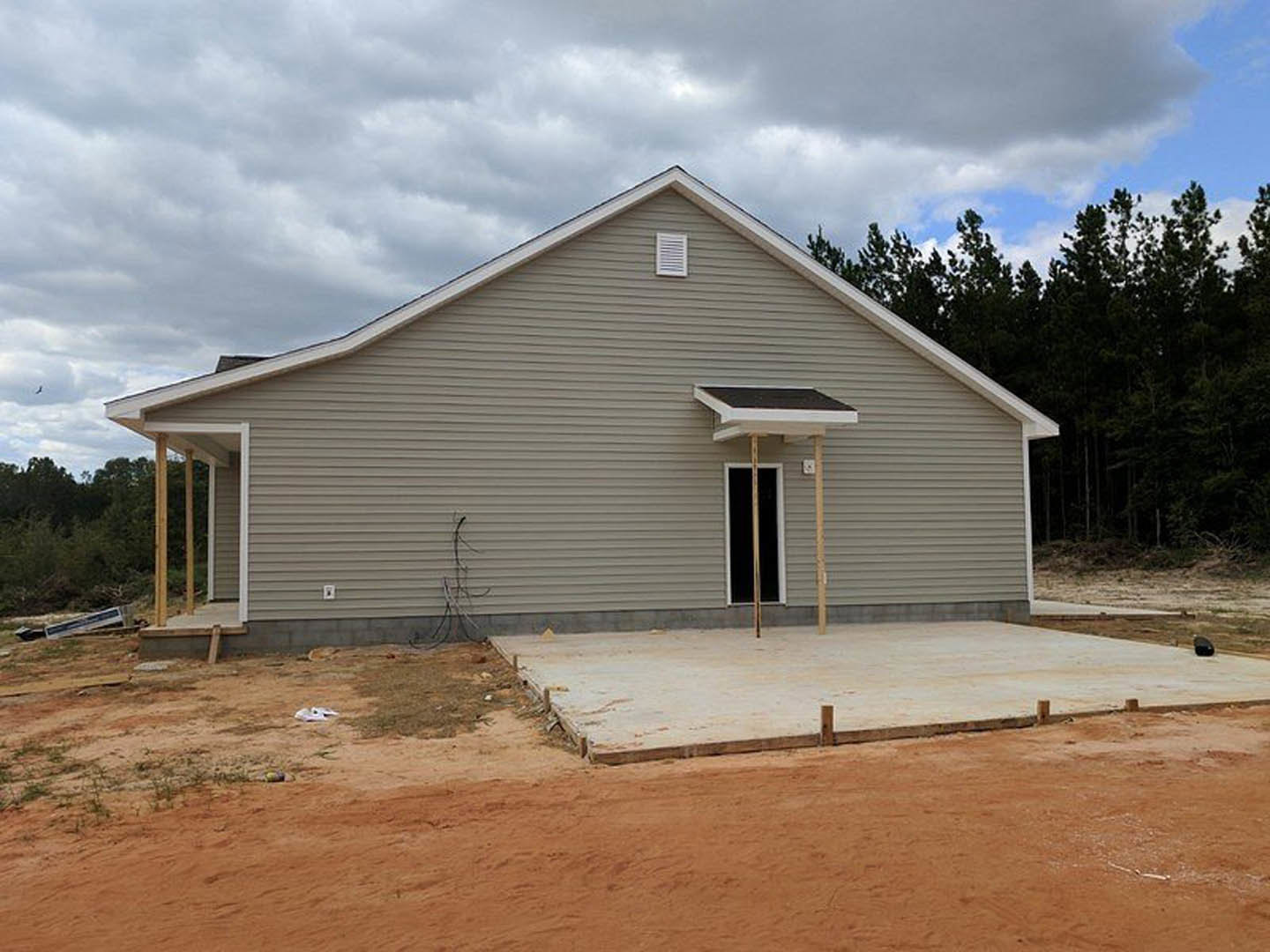 Framed house under construction with exposed wood siding, front porch, and surrounding trees in the background