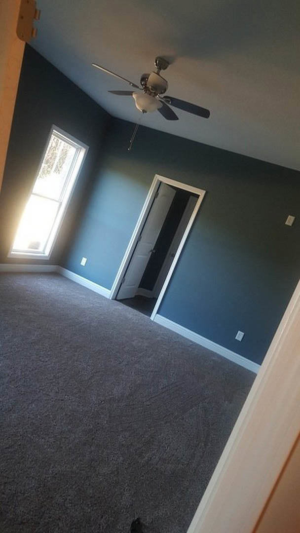 Carpeted room with white door open, ceiling fan with light fixture, window letting in natural light, white walls with molding.